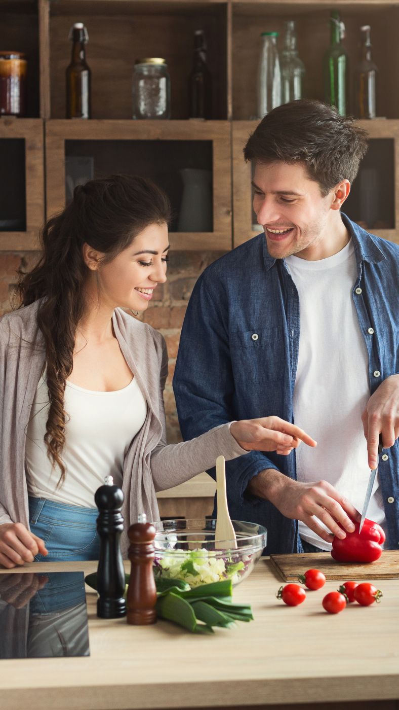 Ein junger Mann und eine junge Frau stehen in einer modernen Küche und bereiten gemeinsam ein Gericht zu. Der Mann schneidet eine rote Paprika auf einem Holzbrett, während die Frau lächelnd auf die Paprika zeigt. Auf der Arbeitsfläche stehen eine Schüssel mit frischem Gemüse, eine Holz-Kochlöffel, Gewürzmühlen, Frühlingszwiebeln und Kirschtomaten. Im Hintergrund sind offene Holzregale mit Gläsern und Flaschen zu sehen.