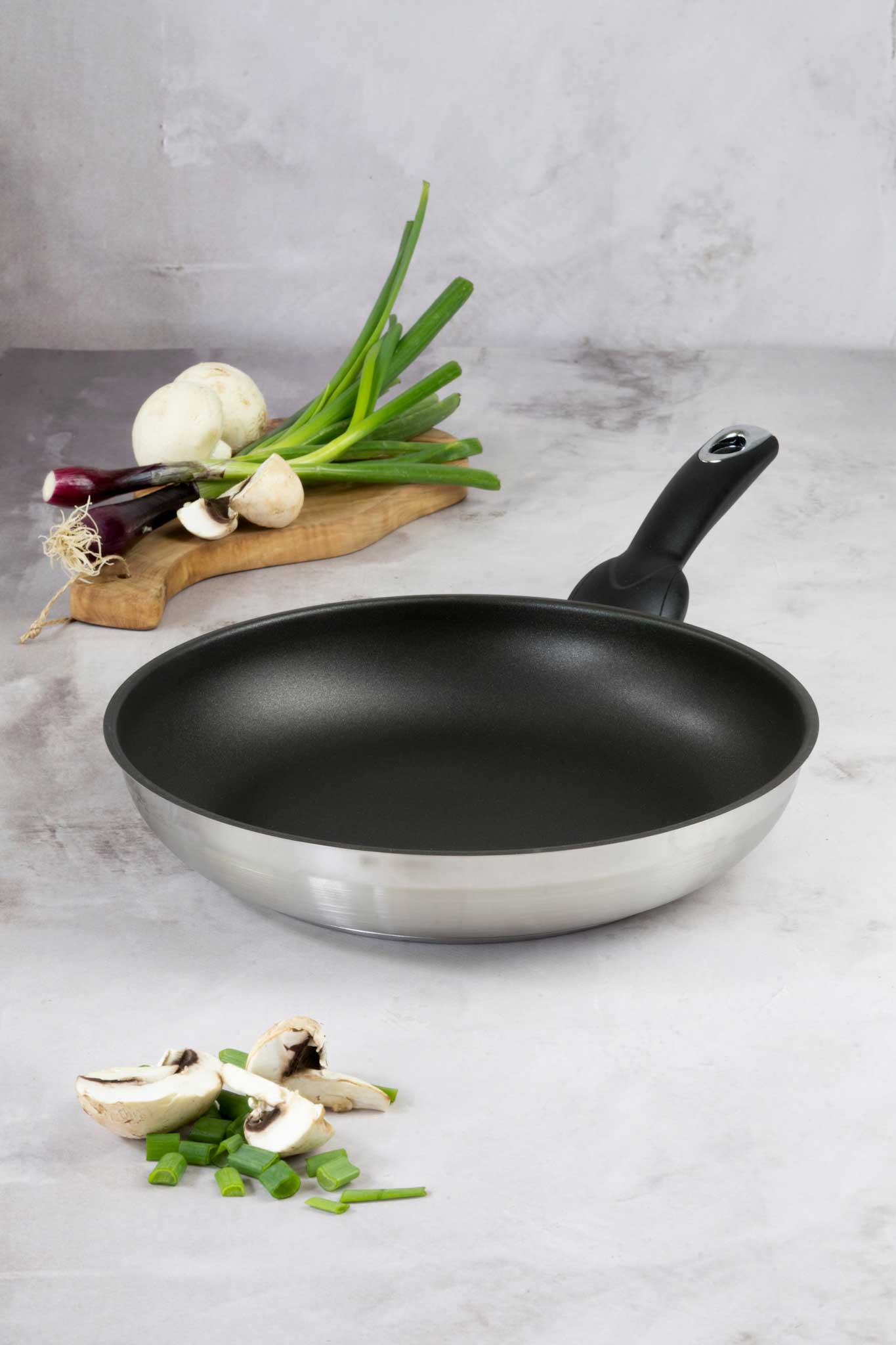 Eine silberne Pfanne mit schwarzer Antihaftbeschichtung steht auf einer hellen Marmorarbeitsfläche. Im Hintergrund liegt auf einem Holzbrett ein Bund Frühlingszwiebeln und einige weiße Zwiebeln. Im Vordergrund befinden sich geschnittene Champignons und Lauchzwiebelringe.