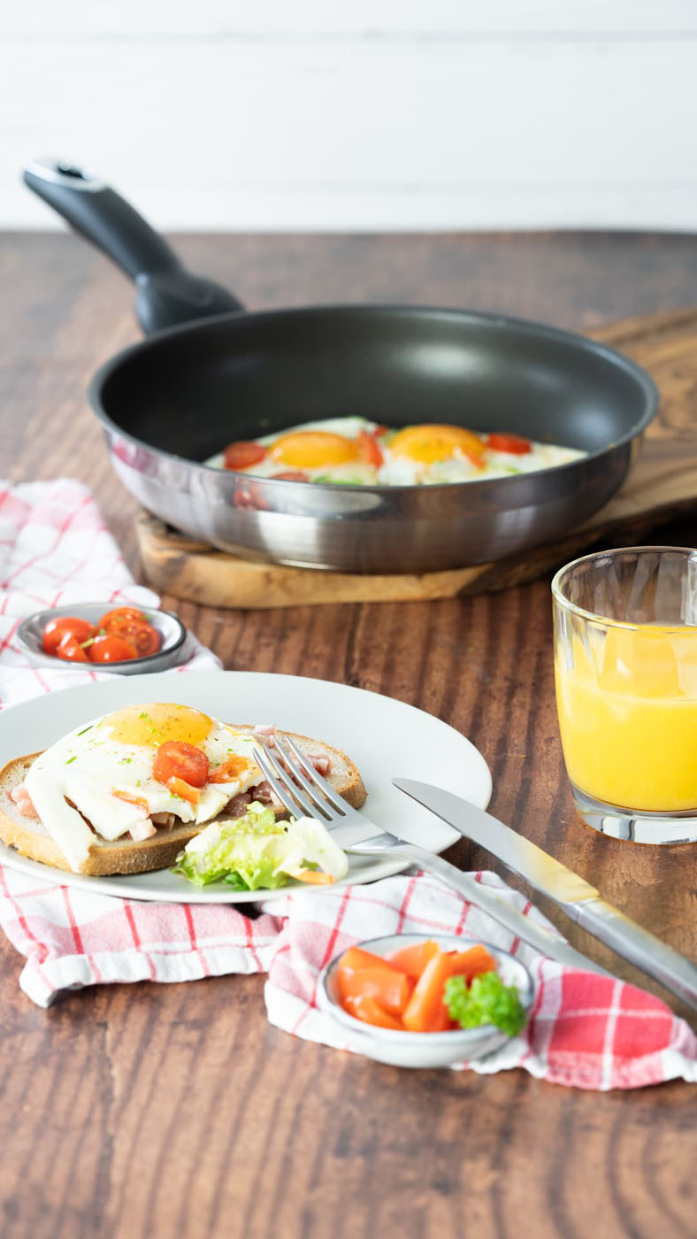 Ein gedeckter Frühstückstisch mit einem Teller, auf dem ein belegtes Brot mit Spiegelei, Schinken, Käse und Tomaten sowie etwas Salat liegt. Daneben stehen Messer und Gabel auf einer rot-weiß karierten Serviette. Im Hintergrund befindet sich eine Pfanne mit zwei Spiegeleiern und Tomaten, daneben ein Glas mit frisch gepresstem Orangensaft. Der Tisch ist aus dunklem Holz.