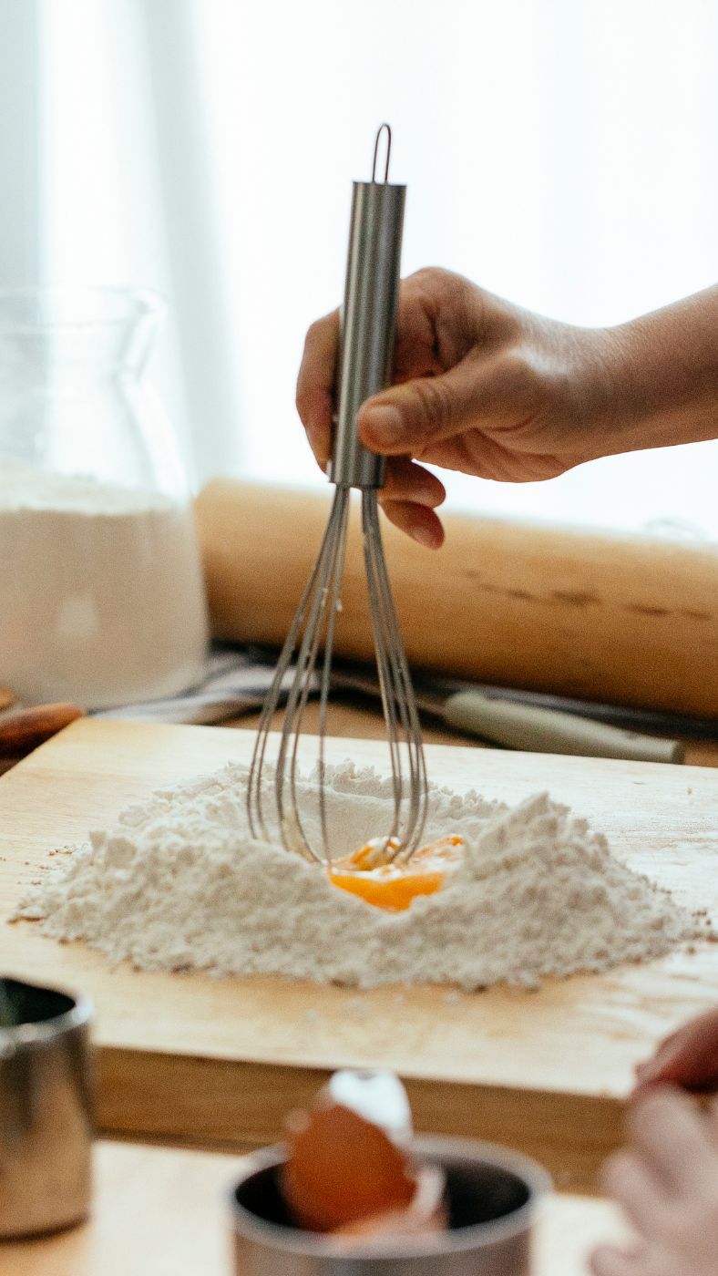 Eine Hand hält einen Schneebesen über eine Mulde aus Mehl auf einem Holzbrett. In der Mulde befindet sich ein aufgeschlagenes Ei. Im Hintergrund stehen ein Nudelholz, ein Glasbehälter mit Mehl und weitere Backutensilien. Im Vordergrund ist eine zerbrochene Eierschale zu sehen.