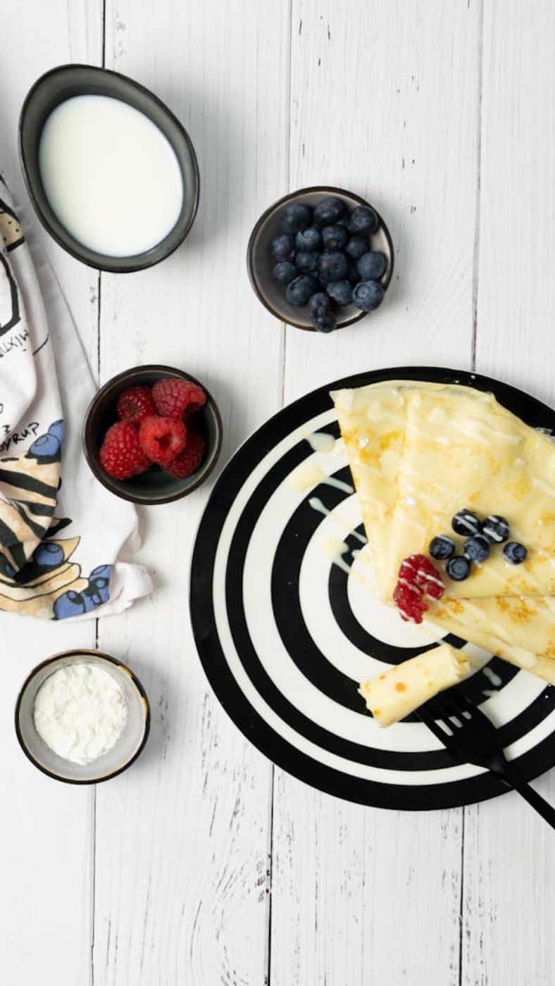 Ein schwarz-weiß gemusterter Teller mit einem zusammengefalteten Crêpe, garniert mit frischen Himbeeren, Blaubeeren und heller Soße. Daneben liegt eine schwarze Gabel. Auf dem weißen Holztisch stehen kleine Schälchen mit Blaubeeren, Himbeeren, Mehl und Milch. Links im Bild liegt ein Geschirrtuch mit einem bunten Muster.