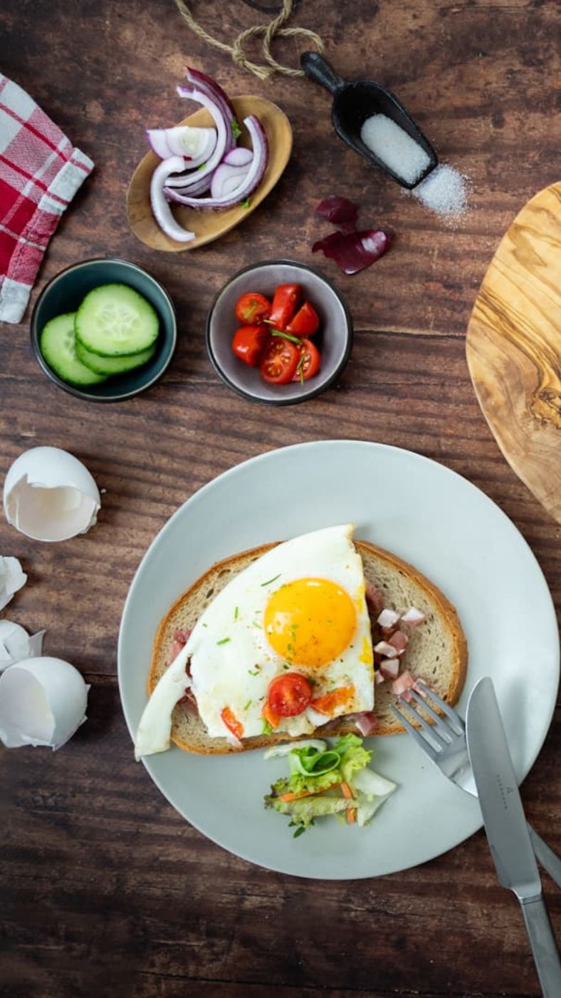 Ein Frühstücksteller mit einer Scheibe Brot, belegt mit gewürfeltem Schinken, einem Spiegelei und halbierten Kirschtomaten. Daneben etwas Blattsalat. Auf dem Holztisch liegen eine Gabel, ein Messer, eine zerbrochene Eierschale, Gurkenscheiben, rote Zwiebelringe, Salz, Kirschtomaten und ein rot-weiß kariertes Küchentuch.
